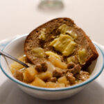 A white and blue plate with a matching bowl. The bowl has a hearty sausage and potato soup with cubed potatoes and small pieces of sausage. There is also a slice of fresh bread topped with a generous amount of butter.