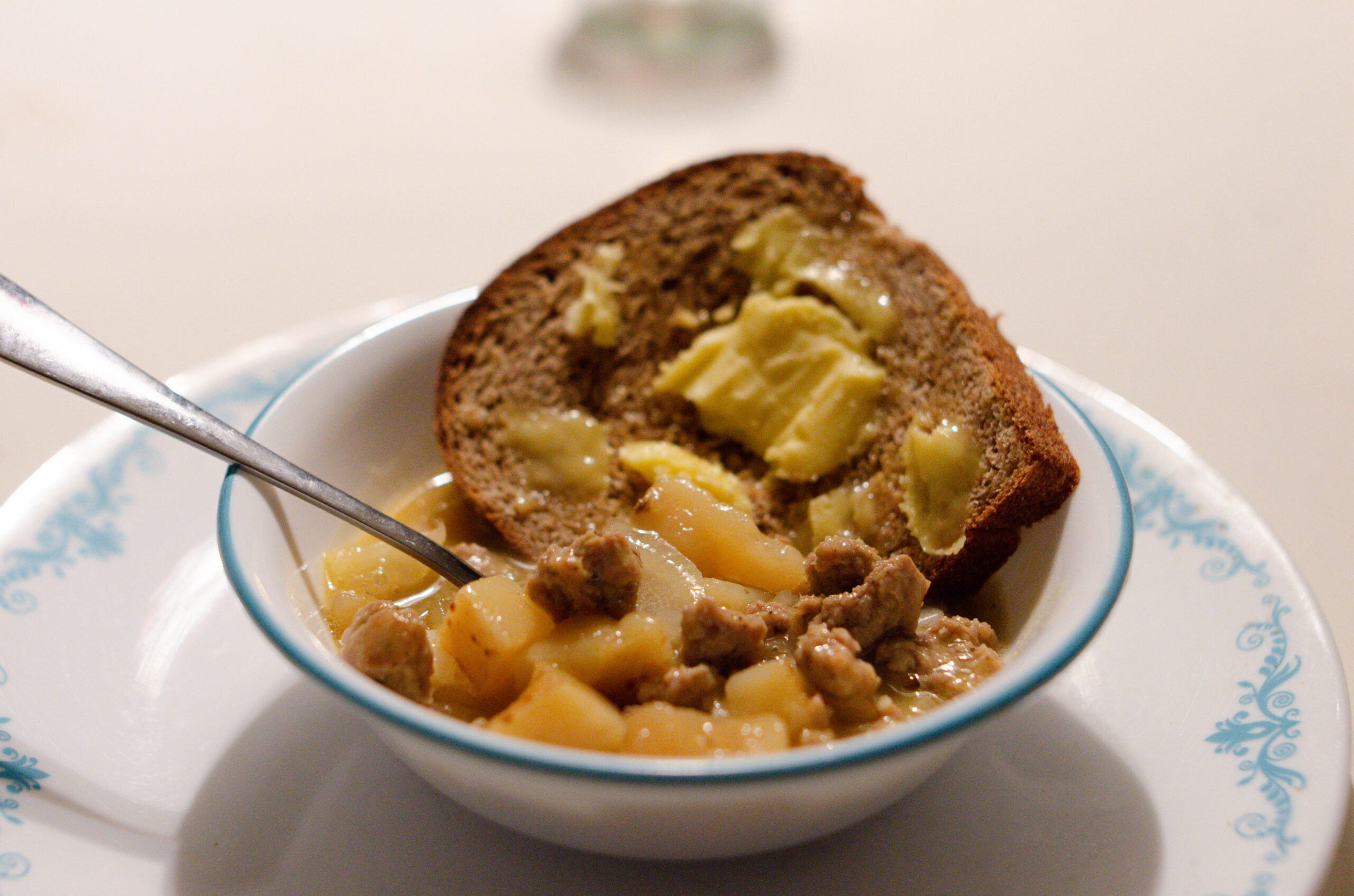 A white and blue plate with a matching bowl. The bowl has a hearty sausage and potato soup with cubed potatoes and small pieces of sausage. There is also a slice of fresh bread topped with a generous amount of butter.